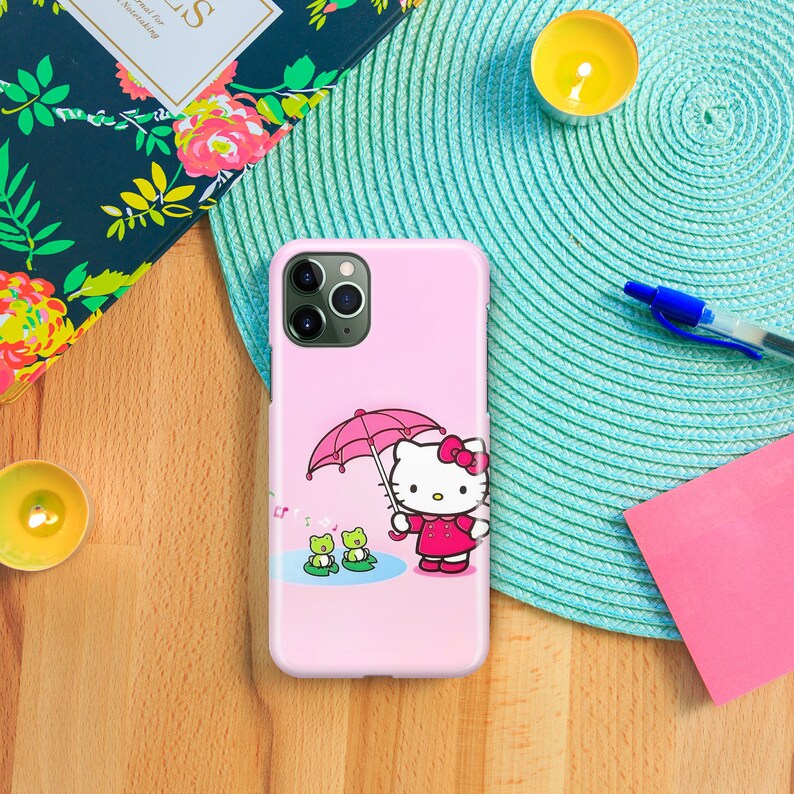 以下が含まれることがあります： ピンクのiPhoneケースにハローキティのデザインが施されています。ハローキティはピンクの傘を持っており、背景には緑色のカエルが2匹います。
