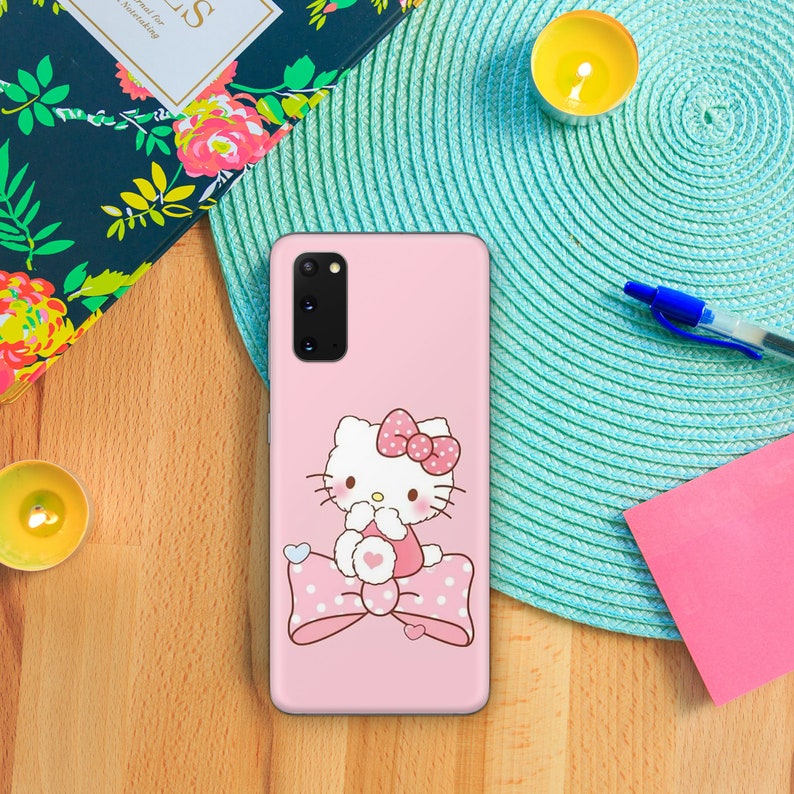 Puede incluir: Una funda de tel&eacute;fono rosa con un personaje Hello Kitty blanco que lleva un lazo rosa y est&aacute; sentado en un lazo rosa con lunares blancos. La funda del tel&eacute;fono tiene una lente de c&aacute;mara negra.