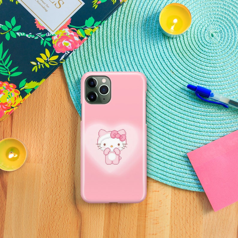 Puede incluir: Una funda rosa para iPhone con un personaje de Hello Kitty en el centro. La funda tiene un recorte transparente para el objetivo de la c&aacute;mara y un fondo rosa.