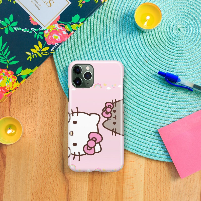 Puede incluir: Una funda de tel&eacute;fono rosa con un Hello Kitty blanco y un gato Pusheen marr&oacute;n. La funda tiene un dise&ntilde;o de arco&iacute;ris y nubes.