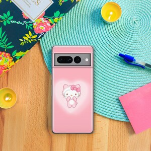 以下が含まれることがあります： ピンクの携帯電話ケースにハローキティのデザインが施されています。ケースは背面が透明で、前面はピンクで、白いハローキティの画像が描かれています。
