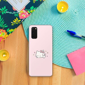 以下が含まれることがあります： ピンクの携帯電話ケースに、ピンクのリボンとキラキラが付いた白いハローキティのキャラクターが描かれています。携帯電話ケースは、Samsung Galaxy S20用です。