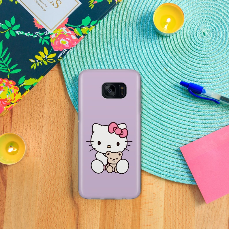 May include: A lavender phone case featuring a white Hello Kitty character holding a brown teddy bear. Hello Kitty has a pink bow on her head.