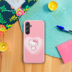 May include: A pink phone case with a Hello Kitty character design. The case has a clear back and a pink Hello Kitty character with a pink bow on her head. The phone case has a camera cutout on the back.
