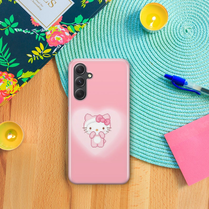 May include: A pink phone case with a Hello Kitty character design. The case has a clear back and a pink Hello Kitty character with a pink bow on her head. The phone case has a camera cutout on the back.