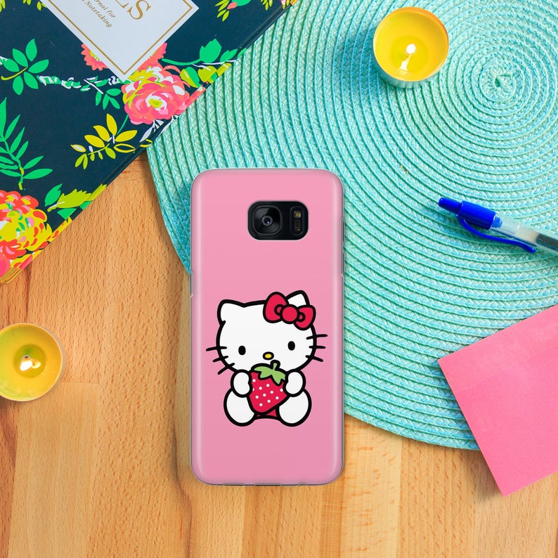 May include: A pink phone case featuring a white Hello Kitty character holding a red strawberry. The case has a white border.
