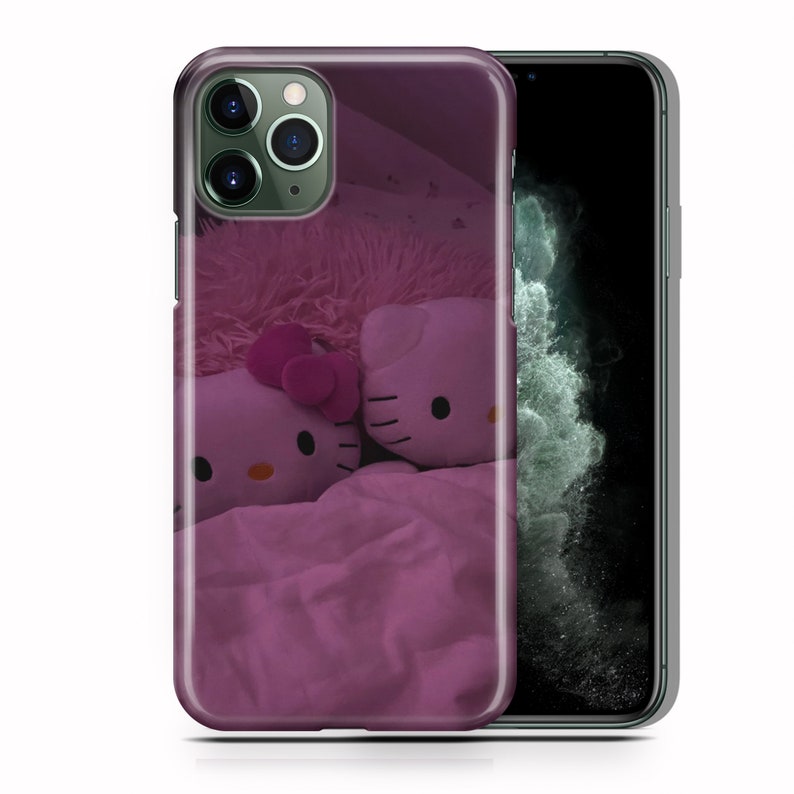 以下が含まれることがあります： ピンクと白のハローキティの携帯電話ケース。ピンクのベッドにハローキティのぬいぐるみが2体置かれています。