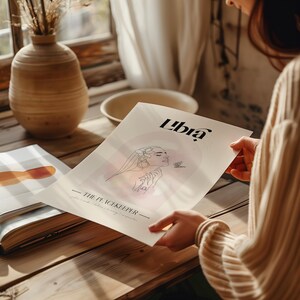 May include: A white poster featuring a line drawing of a woman's face with flowers in her hair. The poster has the text "Libra - The Peacekeeper -" and a butterfly. The poster is on a wooden table.