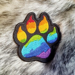 Könnte beinhalten: Gestickter Pfotenabdruck mit Regenbogen. Der Patch ist schwarz mit einem Regenbogenmuster.