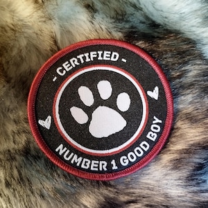 May include: A black and red embroidered patch with a white paw print in the centre. The text "CERTIFIED - NUMBER 1 GOOD BOY" is written around the paw print.