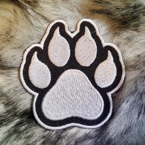 Könnte beinhalten: Ein gestickter Pfotenabdruck-Patch in Schwarz und Weiß. Die Pfote ist weiß mit schwarzen Umrissen und einem schwarzen Rand.