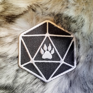 Op de afbeelding: Een geborduurde patch in zwart en wit, gevormd als een twintigzijdige dobbelsteen met een witte voetafdruk in het midden.