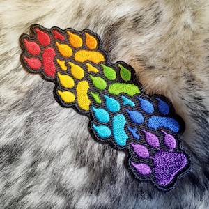 Może przedstawiać: Naszywka z tęczową odbitką łapy z czerwonymi, pomarańczowymi, żółtymi, zielonymi, niebieskimi i fioletowymi odbitkami łap na czarnym tle.