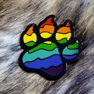 Op de afbeelding: Een zwarte pootafdrukspeld met een regenboogontwerp. De speld heeft de vorm van een pootafdruk, waarbij elke voetzool en teen in de kleuren van de regenboog zijn gekleurd. De speld staat tegen een harige achtergrond.