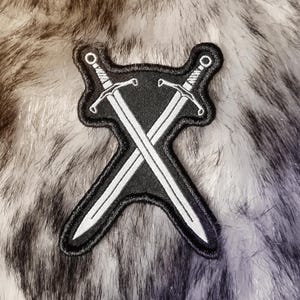 Könnte beinhalten: Ein gestickter schwarzer und weißer Aufnäher mit zwei gekreuzten Schwertern. Die Schwerter sind silberfarben und der Hintergrund ist schwarz.