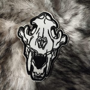 Könnte beinhalten: Gestickter Aufnäher mit einem detaillierten, stilisierten Totenkopf-Design in Schwarz und Weiß. Der Aufnäher hat ein strukturiertes Aussehen, wobei die Merkmale des Schädels vor einem weißen Hintergrund deutlich definiert sind.