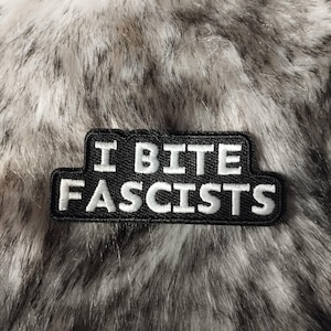 Puede incluir: Un parche rectangular negro con texto bordado en blanco que dice "I BITE FASCISTS". El parche está sobre un fondo de piel sintética en tonos grises y blancos.