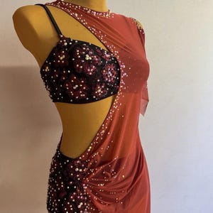 May include: A red dance costume with a black sequined bodice and skirt. The costume features a one-shoulder design with a sheer overlay and is adorned with sparkling embellishments. The garment is displayed on a mannequin.