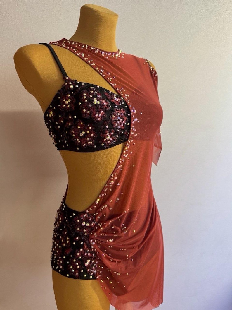 May include: A red dance costume with a black sequined bodice and skirt. The costume features a one-shoulder design with a sheer overlay and is adorned with sparkling embellishments. The garment is displayed on a mannequin.
