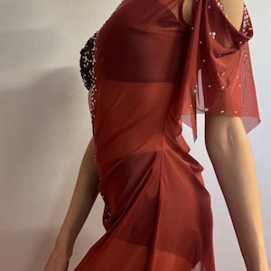 May include: A sheer, rust-colored dress with a flowing design. The dress features a cutout shoulder design and is embellished with small, sparkling beads. The bodice has a black, beaded accent. The dress is a unique piece of apparel.