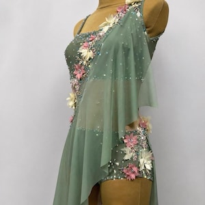 May include: Sage green dance costume with floral embellishments. The one-shoulder leotard features a sheer overlay and is adorned with pink, white, and clear decorative accents. The costume is displayed on a mannequin.