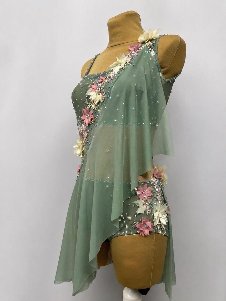 May include: Sage green dance costume with floral embellishments. The one-shoulder leotard features a sheer overlay and is adorned with pink, white, and clear decorative accents. The costume is displayed on a mannequin.