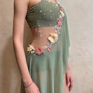 May include: Sage green dance costume with floral embellishments and sparkling accents. The dress features a sheer panel, asymmetrical skirt, and delicate spaghetti straps. Pink and cream-colored flowers adorn the bodice and skirt.