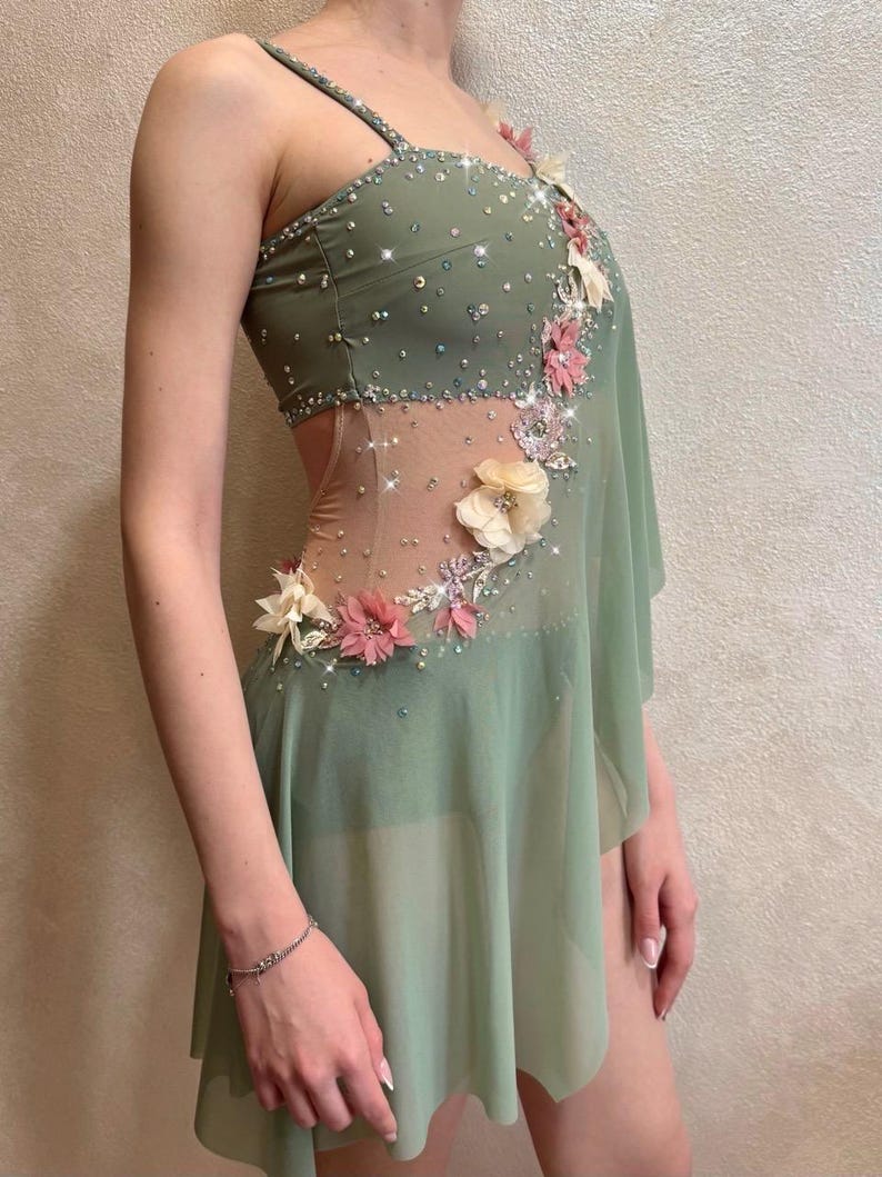 May include: Sage green dance costume with floral embellishments and sparkling accents. The dress features a sheer panel, asymmetrical skirt, and delicate spaghetti straps. Pink and cream-colored flowers adorn the bodice and skirt.