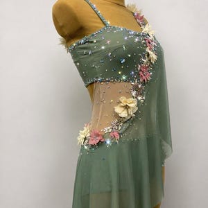 May include: Sage green dance dress with sparkling embellishments and floral accents. The dress features a sheer overlay and delicate straps. The bodice is adorned with crystals and the skirt has a flowing design.