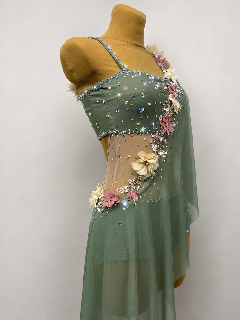 May include: Sage green dance dress with sparkling embellishments and floral accents. The dress features a sheer overlay and delicate straps. The bodice is adorned with crystals and the skirt has a flowing design.