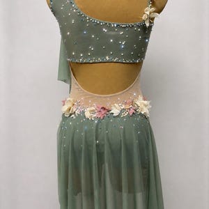 May include: A sage green dance costume with a one-shoulder design and a flowing skirt. The bodice is embellished with sparkling rhinestones and floral accents. The back features a keyhole cutout, and the waistline is adorned with delicate flowers.