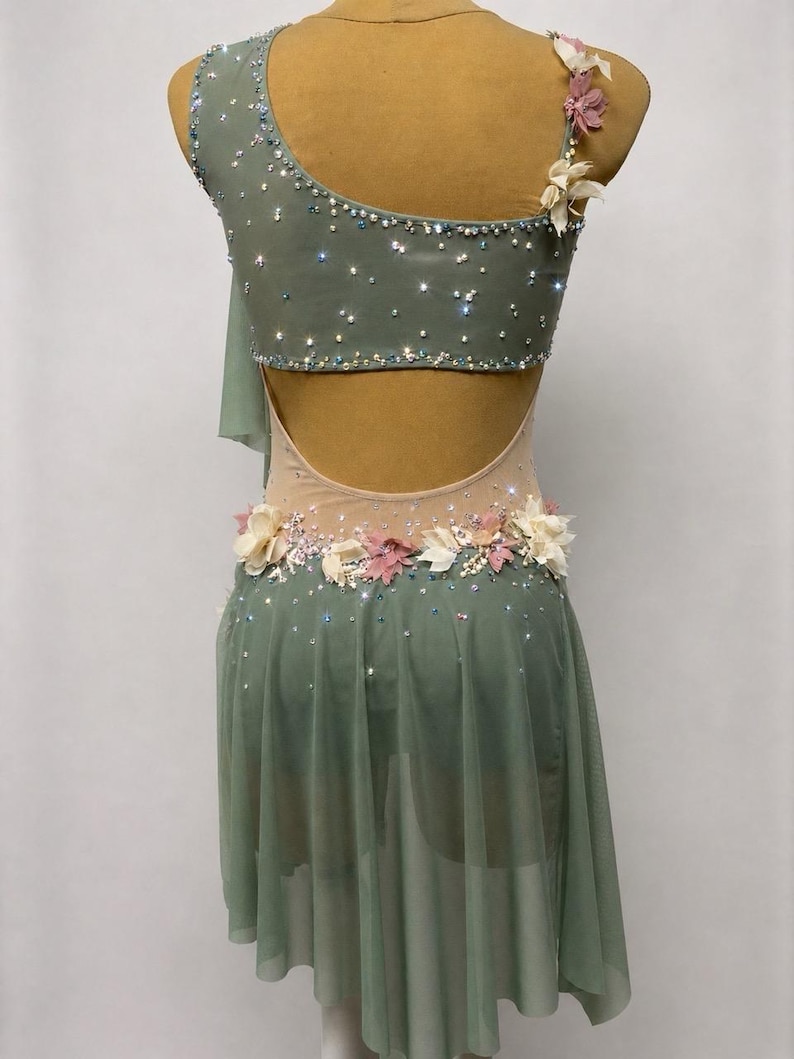 May include: A sage green dance costume with a one-shoulder design and a flowing skirt. The bodice is embellished with sparkling rhinestones and floral accents. The back features a keyhole cutout, and the waistline is adorned with delicate flowers.