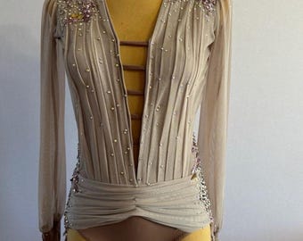 May include: A beige dance leotard with long sleeves and a plunging neckline. The leotard is embellished with rhinestones and sequins. The sleeves are sheer and have a ruffled detail at the cuffs.