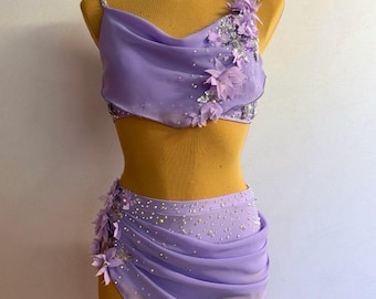 May include: A lavender dance costume set on a mannequin. The set includes a crop top with floral and sequin embellishments and a draped skirt. The costume is made of a sheer fabric and is designed for dance performances.