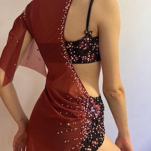May include: A burgundy dance costume with a sheer, asymmetrical overlay adorned with scattered rhinestones. The costume includes a black and burgundy sequined bra and shorts set, creating a glamorous and eye-catching ensemble.