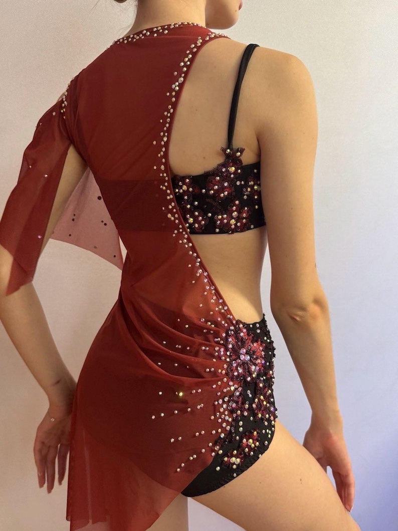 May include: A burgundy dance costume with a sheer, asymmetrical overlay adorned with scattered rhinestones. The costume includes a black and burgundy sequined bra and shorts set, creating a glamorous and eye-catching ensemble.