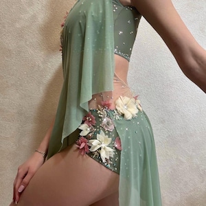 May include: A light green dance costume with floral embellishments. The costume features a leotard base with a sheer overlay and is adorned with white, pink, and cream-colored flowers and sparkling rhinestones. The design includes a flowing cape-like element.