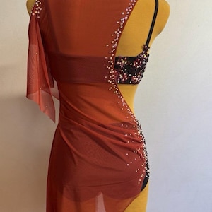 May include: A red dance costume with a sheer overlay and sparkling embellishments. The costume features a one-shoulder design and a black bra top with decorative beading. The fabric drapes elegantly, creating a dynamic silhouette.