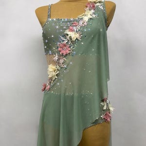 May include: Sage green dance costume with floral embellishments. The asymmetrical dress features a sheer overlay, sparkling accents, and delicate pink and cream-colored fabric flowers. The bodice is adorned with shimmering rhinestones.