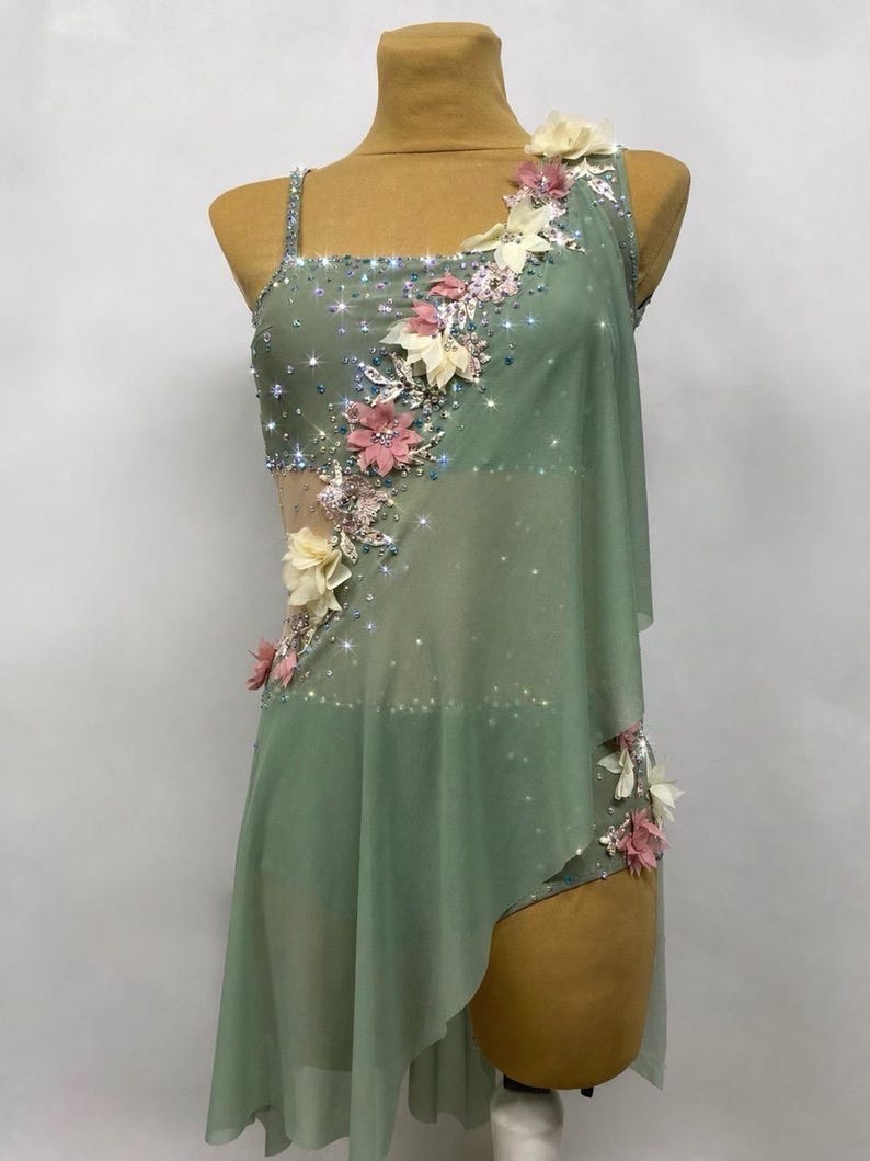 May include: Sage green dance costume with floral embellishments. The asymmetrical dress features a sheer overlay, sparkling accents, and delicate pink and cream-colored fabric flowers. The bodice is adorned with shimmering rhinestones.