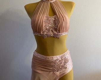 May include: A blush-colored dance costume with a halter top, a skirt with a ruffled hem, and delicate floral embroidery. The costume is adorned with rhinestones.