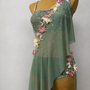 May include: Sage green dance costume with floral embellishments. The one-shoulder leotard features a sheer overlay and is adorned with pink, cream, and crystal accents. The costume is displayed on a mannequin.