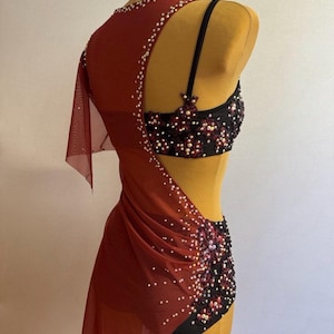 May include: A burgundy dance costume with a black and red embellished bodice and shorts. The costume features a sheer, draped overlay adorned with white beads. The backless design is displayed on a mannequin.