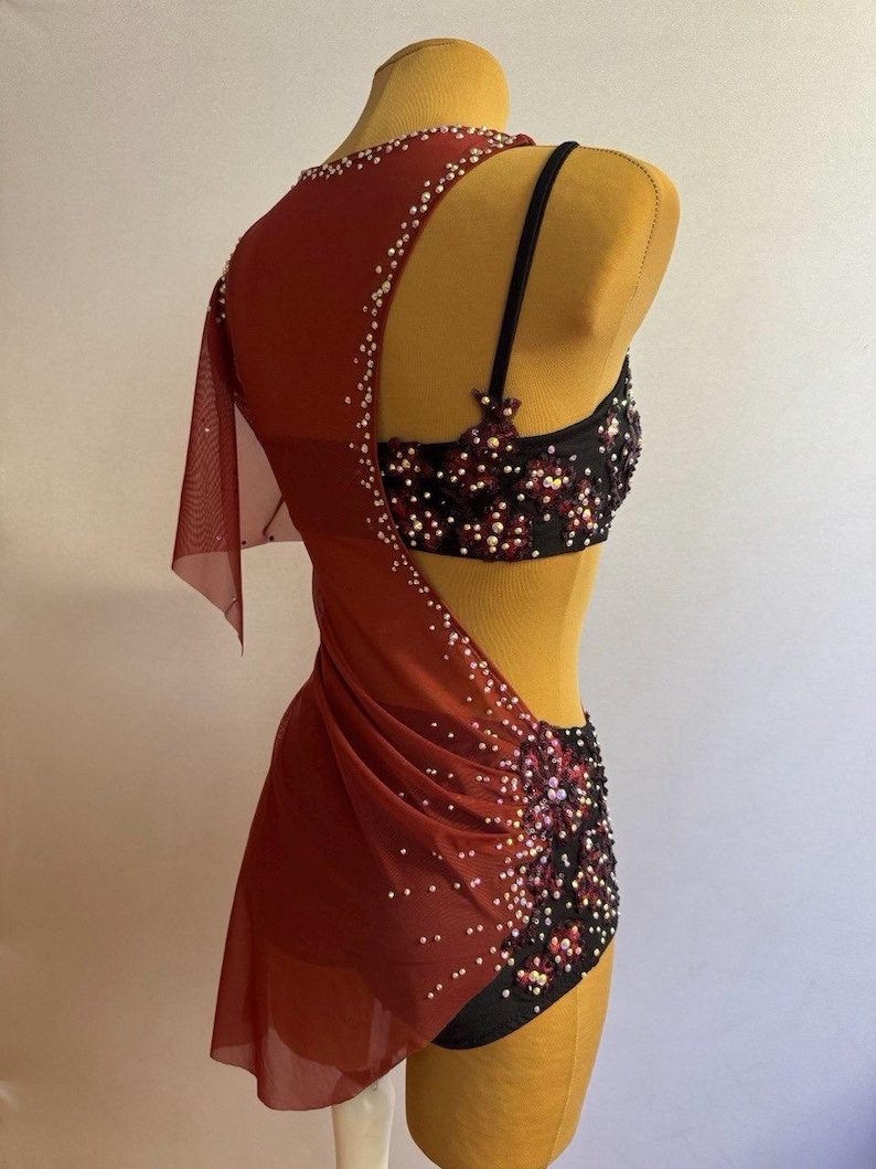 May include: A burgundy dance costume with a black and red embellished bodice and shorts. The costume features a sheer, draped overlay adorned with white beads. The backless design is displayed on a mannequin.