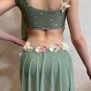 May include: A sage green dance costume with a one-shoulder design. The bodice is embellished with sparkling rhinestones and floral accents. The skirt is a flowing, sheer fabric. The back features a cutout design.