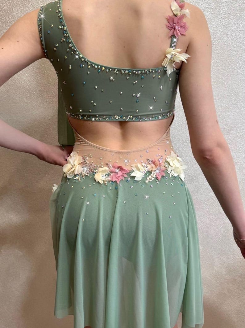 May include: A sage green dance costume with a one-shoulder design. The bodice is embellished with sparkling rhinestones and floral accents. The skirt is a flowing, sheer fabric. The back features a cutout design.