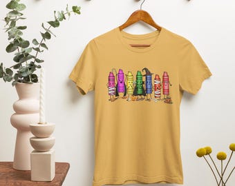 Könnte beinhalten: Senfgelbes T-Shirt mit einer Cartoon-Illustration von anthropomorphen Buntstiften zum Thema Halloween. Die Buntstifte sind in verschiedenen Farben gehalten, darunter Lila, Grün und Blau. Das T-Shirt hängt an einem Holzbügel.