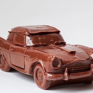 Könnte beinhalten: Ein Schokoladen-Automodell, eine Nachbildung eines klassischen Autos, mit brauner Oberfläche. Das Auto ist detailliert mit einer Windschutzscheibe, Scheinwerfern und einem Kühlergrill.