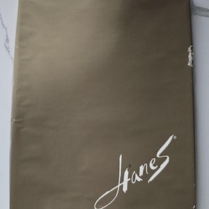 Op de afbeelding: Een rechthoekig pakket verpakt in bruin papier. Het woord "Hanes" is in een wit, cursief schrift in de rechter benedenhoek geschreven. Het pakket is verzegeld met doorzichtige tape.
