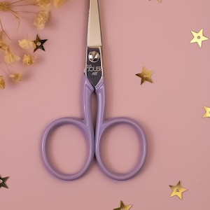 May include: A pair of small, silver-bladed scissors with lavender handles. The scissors have a rounded handle design and the text "BOUER ART" is printed on the blade joint. The background is pink with gold star-shaped confetti and dried flowers.